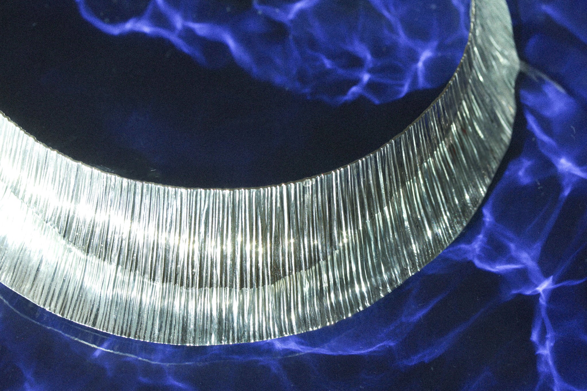 Sculptural silver choker necklace with textured surface, inspired by waterfall flow, shown against a deep blue water-like background with light reflections.